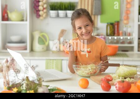 Petite fille en train de faire de la salade sur la cuisine et en utilisant un ordinateur portable Banque D'Images