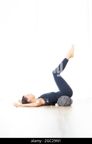 Yoga réparateur avec un traversin. Jeune femme sportive attrayante dans un studio de yoga blanc brillant, allongé sur un coussin de traversin, s'étirant et se détendant pendant Banque D'Images