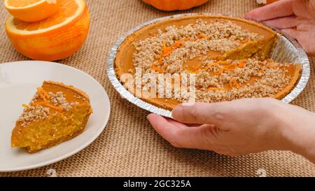 Tarte à la citrouille maison fraîchement cuite décorée de noix écrasées et de zeste d'orange. Femme mains servant la tarte sur une assiette, vue rapprochée Banque D'Images