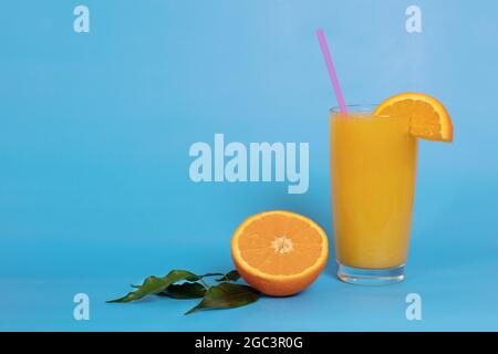 Jus d'orange dans un verre avec paille, décoré d'une tranche d'orange, d'une moitié d'orange avec des feuilles sur fond bleu. Boisson d'été. Copier l'espace Banque D'Images