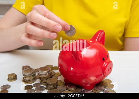 Une main femelle met des moents dans la fente d'une banque de porc rouge, sur la table sont beaucoup de pièces de différents pays. Concept d'enregistrement. Banque D'Images