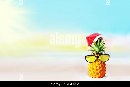 Joli ananas dans des lunettes de soleil miroirs élégantes sur le sable contre l'eau de mer. Porter un chapeau de noël. Concept de Noël et de nouvel an sur un bea tropical Banque D'Images