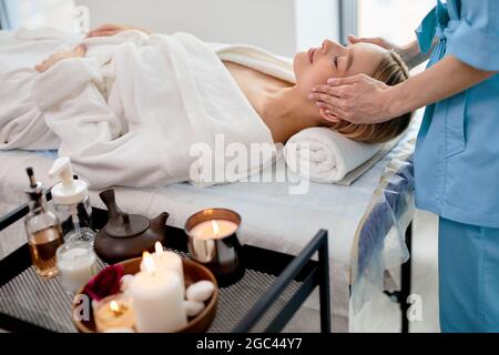 Masseuse féminine non reconnaissable en bleu faisant un massage relaxant à la femme cliente allongée sur le canapé, la femme caucasienne est détendue, profitant de soins spa dans Banque D'Images