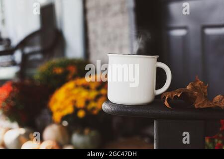 Tasse de café à la vapeur assise sur le bras de la chaise à bascule sur un porche avant qui a été décoré pour l'automne avec des citrouilles et des mamans. Banque D'Images