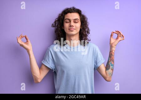 Calme caucasien curly mâle garder calme, méditant isolé en studio sur fond violet. Un homme charmant garde les yeux fermés, engagé dans le yoga Banque D'Images