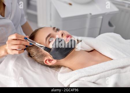 Mains de la femme cosmétologue caucasienne appliquant un masque facial sur le beau visage de la jeune femme caucasienne couché sur le lit de massage dans peignoir, vue latérale po Banque D'Images