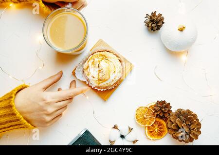 Vue du dessus des mains des femmes touchant le gobelet à café et le tartelet. Fille en pull tricoté est de prendre le petit déjeuner. Dessert sucré, boisson, cône de pin et décorations sur table blanche. Esthétique de la vie. Banque D'Images