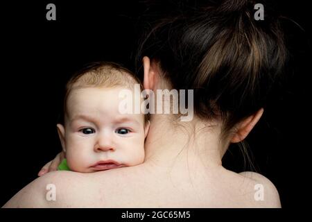 bébé sur l'épaule de maman, regardant l'appareil photo, sans émotion. Sur fond noir. Banque D'Images