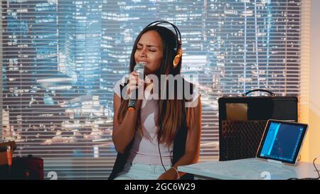 Élégante jeune fille de race mixte chantant dans le microphone à la répétition dans le studio à la maison avec des gratte-ciel en arrière-plan. Banque D'Images