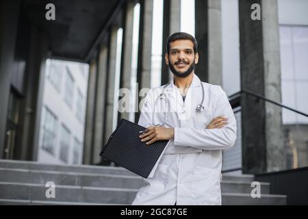Médecin arabe confiant et fiable, en manteau blanc et avec stéthoscope, tenant le bloc-notes et le stylo, debout avec les bras croisés sur l'arrière-plan de l'hôpital moderne. Banque D'Images
