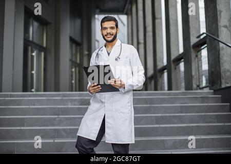 Vue rapprochée de l'avant d'un agréable jeune médecin ethnique du Moyen-Orient en robe blanche, tenant un dossier, debout sur les escaliers de la clinique moderne à l'extérieur, souriant et regardant la caméra Banque D'Images