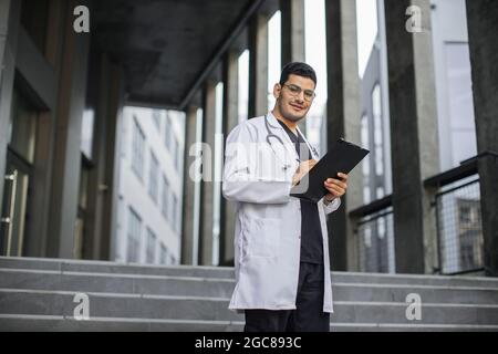 Jeune médecin arabe, descendant les escaliers à l'extérieur de l'hôpital moderne, regardant la caméra et prenant des notes dossier du patient. Médecin marchant à l'extérieur près de l'hôpital et pensant au travail Banque D'Images