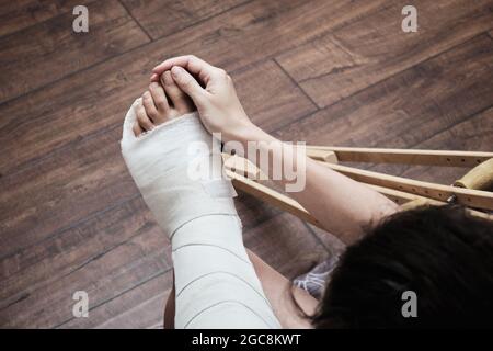 Une femme masse les orteils d'une jambe cassée avec ses mains. Vue de dessus d'une jambe cassée dans un plâtre et des béquilles. Réhabilitation à domicile après une rupture Banque D'Images