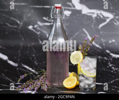 Bouteille en verre avec bouchon et limonade lavande sur fond de marbre noir. Eau aromatique avec citron et glaçons. Banque D'Images
