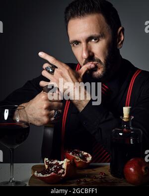 Homme brutal en chemise noire et bretelles à la table avec du jus de grenade frais, tient le couteau et léche son doigt Banque D'Images