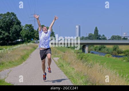 Joyeux et exubérant homme d'âge moyen qui fait du jogging le long d'une rivière en célébrant sa bonne santé et son temps ensoleillé, levant les bras dans l'enthousiasme Banque D'Images