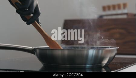 homme main dans des gants en déplaçant quelque chose avec une spatule en bois dans un poêle à frire, photo large Banque D'Images