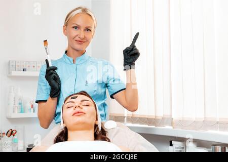 Salon professionnel de cosmétologie. Cosmetolog en gants de latex tenant une brosse de masque et montrer avec l'index sur le côté droit. Le client se trouve sur la procédure. Banque D'Images