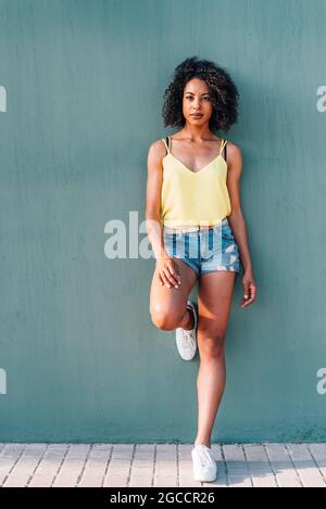 portrait vertical d'une femme africaine avec un fond de mur vert. Elle se penche sur le mur vert avec des vêtements d'été et regarde l'appareil photo Banque D'Images