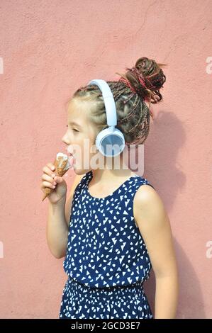 Petite fille tressée à l'écoute de musique portant des écouteurs et de manger de glace. Des vibes d'été. Banque D'Images