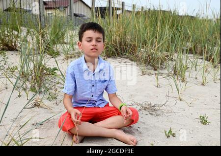 Un beau garçon d'école en short orange et en chemise bleue est assis sur le sol sablonneux dans une position lotus et médite calmement sur le fond de la nature. Banque D'Images