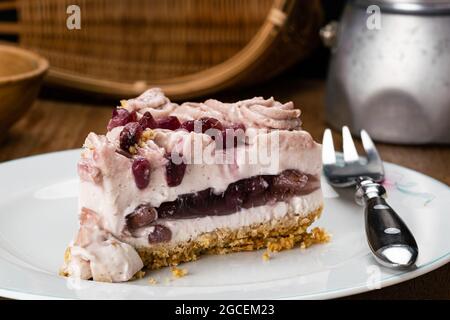 Gros plan de cheesecake maison à la grenade avec fourchette en métal dans un plat en céramique blanche avec une tasse en bois, un panier en bambou et une cafetière en moka Banque D'Images