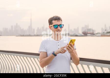 Un jeune homme tape sur un smartphone ou utilise une application dans la rue sur fond de coucher de soleil et du canal de Dubai Creek et du puits de ciel de Burj Khalifa Banque D'Images