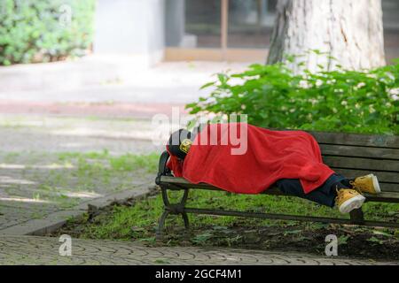 Saint John, N.-B., Canada - le 24 juillet 2021 : personne dormant sur un banc de parc. Ils utilisent une serviette rouge comme couverture. Face non visible. Banque D'Images