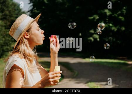 Jolie femme blonde soufflant des bulles de savon. Magnifique parc verdoyant ou forêt en arrière-plan - vue latérale Banque D'Images