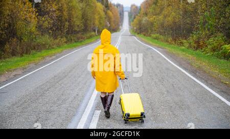 Une femme en imperméable jaune marche le long de la route avec une valise jaune en automne Banque D'Images