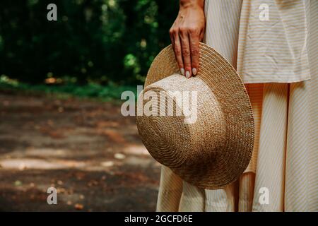 Photo en gros plan de la main d'une femme tenant un chapeau de paille sur le fond d'une robe blanche. Forêt de fées en arrière-plan Banque D'Images