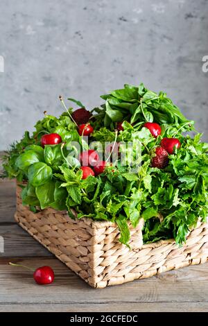 Diverses herbes parfumées fraîches avec cerises et fraises dans un panier en osier sur fond de bois. Concept d'alimentation saine. Récolte d'été dans les jardins. Banque D'Images