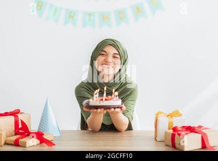 Jeune fille musulmane dans hijab tenant un délicieux gâteau d'anniversaire à la table avec des boîtes cadeaux, célébrant des occasions festives à la maison. Adolescent islamique ayant yu Banque D'Images