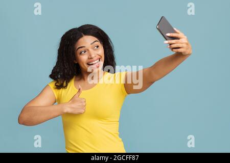 Une femme afro-américaine excitée prend le selfie sur son smartphone et montre le pouce vers le haut sur fond bleu. Selfie sur l'appareil, blogger et la vidéo de tournage pour Banque D'Images