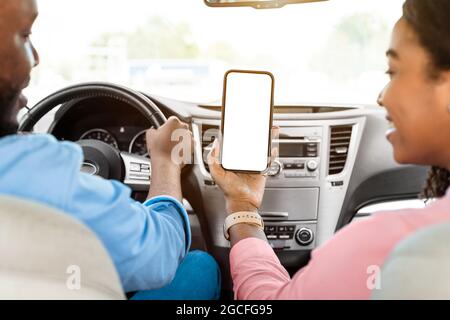 Vue arrière au-dessus de l'épaule d'une femme heureuse noire tenant un smartphone avec écran blanc vierge, maquette, assise sur le siège avant en voiture. Co Banque D'Images