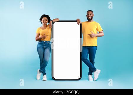 Excellente application mobile. Couple afro-américain pointant vers un smartphone géant avec maquette, faisant la promotion d'une application ou d'un site Web, d'un produit ou d'un service publicitaire, Banque D'Images