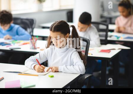 Une mienne mignonne assise à un bureau dans une salle de classe à l'école élémentaire. Fille étudiante faisant le test à l'école primaire. Les enfants écrivent des notes pendant le Banque D'Images