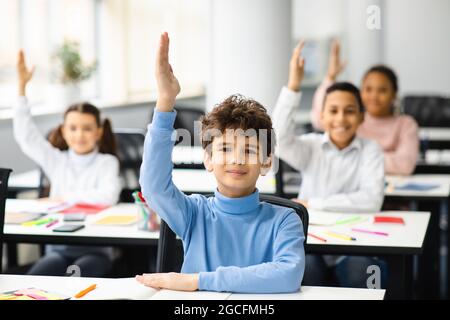 Premier jour d'école. Portrait d'un mignon garçon assis à table en classe, groupe de petits élèves multiculturels qui se soulèvent tous les mains pour une réponse, étudier Banque D'Images