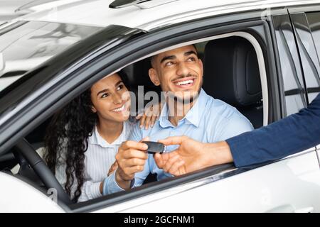 Un couple du Moyen-Orient heureux qui achète une voiture dans une immense salle d'exposition, prend la clé d'une voiture chez un jeune vendeur élégant tout en étant assis à l'intérieur de la voiture. Joyeux arabe m Banque D'Images