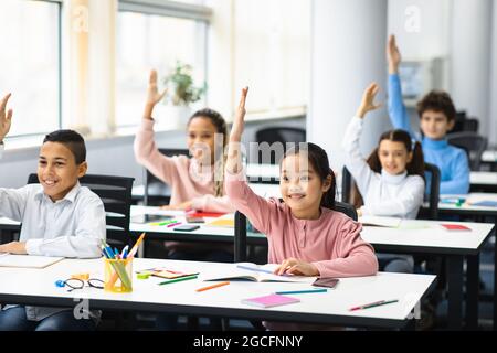 Retour à l'école. Multiculturel Groupe diversifié de petits écoliers de l'élémentaire heureux assis à des bureaux dans la salle de classe, levant les mains pour un an Banque D'Images