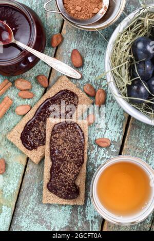 Confiture de prunes maison dans un pot en verre et sur des craquelins de seigle servis avec des fèves de cacao crues, du thé et des prunes fraîches sur du foin sec sur la table de la turqouoise ancienne au-dessus de la vue verticale Banque D'Images