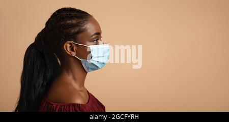 Vue latérale d'une jeune femme noire dans un masque de protection regardant l'espace de copie pour le texte ou la publicité sur fond beige studio, portrait de gros plan, Banque D'Images