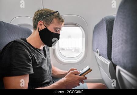 Homme de voyage portant un masque facial pendant le vol en utilisant le téléphone dans l'avion. Jeune homme d'affaires en noir tenant le téléphone portable pour la preuve de passeport de vaccin pendant Banque D'Images