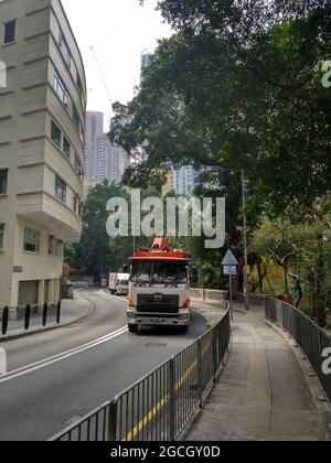 Une rue et un vieux trottoir à Hong Kong avec un grand camion de nettoyage ou de déplacement à côté d'un bâtiment moderne brillant et des arbres verts luxuriants. Banque D'Images