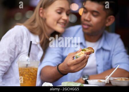 Jeune couple multi-ethnique appréciant de délicieux rouleaux de printemps avec des sauces dans le café extérieur Banque D'Images