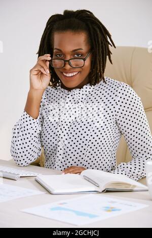 Jeune entrepreneur positif en chemisier à pois travaillant avec des documents au bureau, en ajustant des lunettes et en souriant à l'appareil photo Banque D'Images