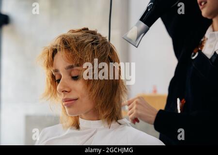 Coiffeur asiatique jeune homme portant un tablier séchant les cheveux de jeune homme beau dans un salon de coiffure moderne tout en se concentrant sur le travail Banque D'Images