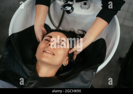 Vue en grand angle de la belle jeune femme asiatique se détendant avec les yeux fermés avec la tête dans le lavabo, tandis que les mains de coiffeuse massant les cheveux et les points de pression pressants Banque D'Images