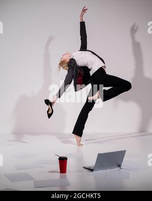 Une ballerine pieds nus gracieuse dans un costume d'affaires sautant avec des chaussures dans ses mains sur un fond blanc. Banque D'Images