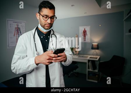 Infirmière masculine de race mixte debout dans le bureau de médecins textant sur un appareil cellulaire Banque D'Images